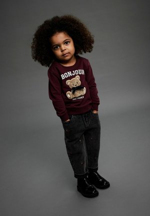 Weinrote Sweatshirt mit Teddy-Bär-Grafik, kombiniert mit schwarzen bestickten Jeans. Glänzende schwarze Knöchelstiefel runden das Outfit ab.