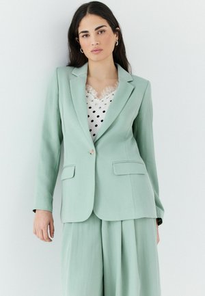 Woman with long dark hair wearing a light green blazer and wide-leg pants over a white polka-dot lace-trimmed top, standing against a white wall.