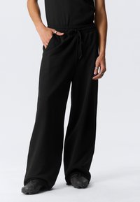 Άτομο που φοράει φαρδιά μαύρη παντελόνια με κορδόνι στη μέση, μαύρα παπούτσια με υφασμένα πέλματα, μαύρο τοπ και πολλά ασημένια δαχτυλίδια στα δάχ τυλα.