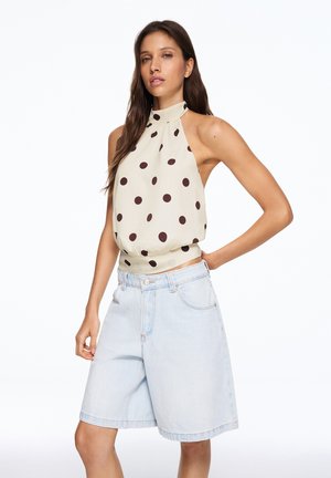 Vrouw draagt een beige mouwloze haltertop met grote bruine stippen en lichtblauwe denim knielange korte broek.
