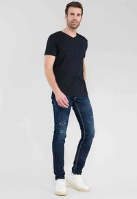 Zwart V-hals T-shirt gecombineerd met donkerblauwe slim-fit jeans met distressed accenten en witte sneakers, allemaal tegen een neutrale achtergrond.
