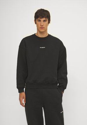 Sweatshirt noir avec un col rond et des épaules tombantes. Présente un petit logo blanc à l'avant. Le matériau semble doux et texturé.