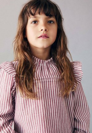 Bambina con lunghi capelli castani e occhi espressivi, che indossa una blusa a righe rosa con maniche a volant, su uno sfondo neutro.