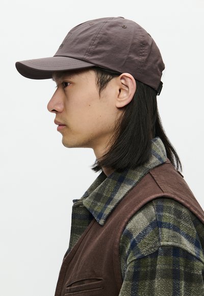 Jaunas vyras su tiesiais juodais plaukais, dėvintis rudą kepurę, margą marškinėlį ir rudą liemenę, besisukantis į kairę priešais paprastą baltą foną.