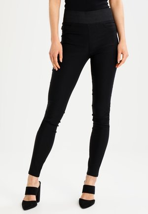 Mujer con pantalones pitillo negros de talle alto y mules negros de tacón con tiras anchas, de pie sobre fondo blanco.