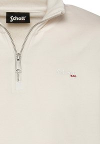 Kremowa bluza z zamkiem błyskawicznym, wykonana z miękkiego materiału. Posiada wysoki kołnierz, srebrny zamek oraz haftowane logo "Schott NYC" w bieli i czerwieni.
