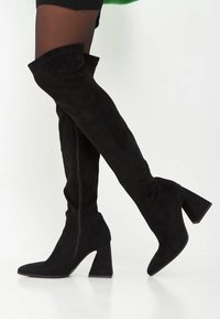 Schwarze Wildleder-Stiefel über das Knie haben eine spitze Zehe, einen klobigen Absatz und einen seitlichen Reißverschluss für einfaches Anziehen. Glatte Textur ohne sichtbare Muster.