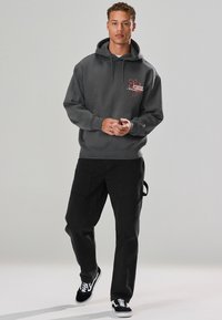 Felpa grigia con tasca frontale e testo grafico rosso, abbinata a pantaloni cargo neri. Il modello posa in modo casual, indossando sneakers nere.