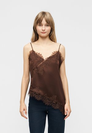 Joven mujer de pie con una camiseta de seda marrón con detalles de encaje y jeans azul oscuro, sobre un fondo claro y sencillo.