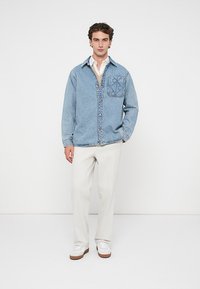Camisa de denim azul clara com botão à frente, um bolso de remendo no lado esquerdo do peito, combinada com calças de cor creme e ténis brancos.