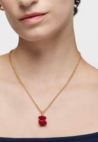 Collar de cadena dorada con un colgante de un osito de peluche rojo, con cabeza y cuerpo redondeados, que cuelga de una cadena de eslabones simple.