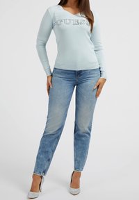 Suéter de manga larga azul claro con la palabra "GUESS" en adornos de cristal, combinado con jeans de pierna recta en azul desgastado y tacones altos plateados.