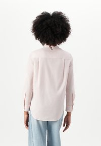 Camicia a maniche lunghe rosa chiaro con orlo arrotondato, spacco posteriore e chiusure a bottone al collo. Tessuto morbido con una texture liscia.