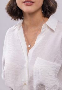 Weißes Hemd mit Knopfleiste und zerknitterter Textur, versehen mit einer Tasche und einer silbernen Halskette mit ovalem Anhänger, der auf einem nackten Hals ruht.