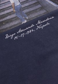Marineblauwe katoenstof met een afgedrukte afbeelding van Diego Maradona op een trap, met daaronder in een cursieve lettertype zijn naam en datum in witte tekst.