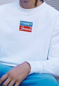 Bílá mikina s vyšitým logem "TOMMY Jeans" v červené a modré barvě. Má žebrované manžety a výstřih. Nositelná s modrými kalhotami.