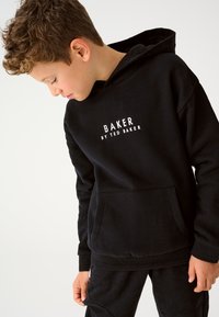 Mustast puuvillasegust kapuutsi ja känguru taskuga dressipluus, millel on eest valged trükitud tähed "BAKER BY TED BAKER". Lõdvestunud istuvus.