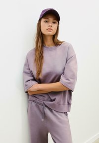 Fioletowy oversize'owy sweter z krótkimi rękawami i okrągłym dekoltem, w zestawie z dopasowanymi spodniami dresowymi i czapką, na jednolitym tle.
