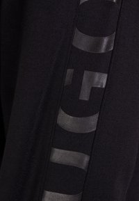 Tessuto nero con un motivo testurizzato e accenti lucidi. Logo visualizzato verticalmente in un nero a contrasto. Morbida texture a coste.