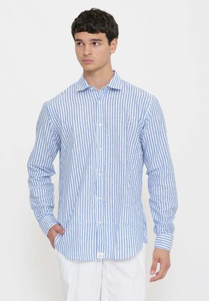 Giovane uomo con capelli corti e scuri che indossa una camicia a righe verticali blu e bianche con bottoni e pantaloni bianchi, che guarda di lato.