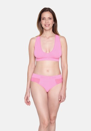 Frau mit schulterlangem Haar, die einen rosa Sport-BH und passende Unterwäsche trägt, steht lächelnd vor weißem Hintergrund.