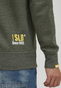 Olijfgroen sweatshirt met ribbelstructuur, beschikt over een gele en witte geborduurde logo aan de linkerkant en een klein geel label op de manchetten.