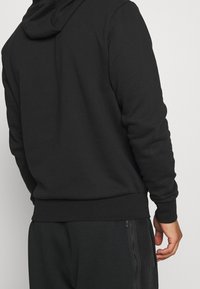 Černá mikina s kapucí a texturovaným povrchem, s žebrovanými manžetami a přímým lemem. Zadní strana má hladký design bez ozdob.