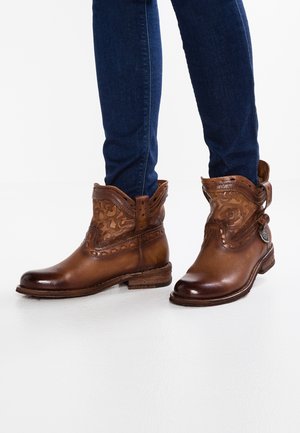 Braune Leder-Stiefeletten mit floraler Prägung, getragen zu dunkelblauen Skinny-Jeans auf weißem Untergrund.