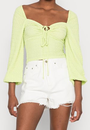 Frau trägt ein limettengrünes, gerafftes Crop-Top mit langen Ärmeln und Frontknoten sowie weiße, ausgefranste, hoch taillierte Jeansshorts.