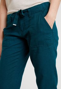 Teal linnbyxor med dragsko i midjan, elastiskt band, sidfickor och avslappnad passform. Har synlig sömnad och en mjuk textur.