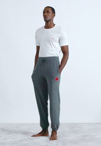 Λευκό κοντομάνικο μπλουζάκι με κόκκινο λογότυπο, συνδυασμένο με γκρι αθλητικό παντελόνι με π�λαϊνή τσέπη και κόκκινη ετικέτα. Ξυπόλητος σε ανοιχτόχρωμη επιφάνεια.