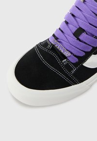 Sneaker in camoscio nero con suola in gomma bianca, lacci viola e brand Vans sulla linguetta. Presenta una striscia laterale bianca e cuciture a contrasto.