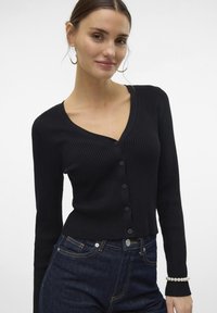 Crna rebrasta cardigan jakna s V-izrezom, zatvaranjem na gumbe i dugim rukavima. Nosi se s tamnim trapericama, a na zapešću ima bisernu narukvicu.