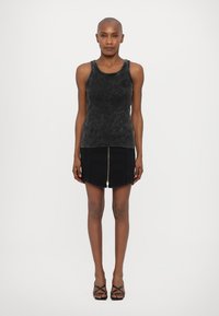 Svart tanktopp med strukturert finish, kombinert med et svart denimskjørt med glidelås foran. Modellen står med hendene på hoftene.