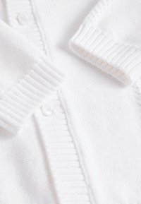 Cardigan blanc en matière douce, avec des poignets côtelés et une fermeture à boutons. Texture lisse avec des accents subtils de motif tricoté.