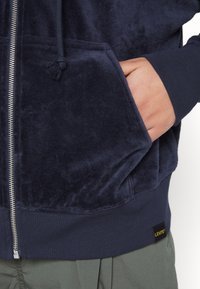 Main dans la poche avant d'un sweat à capuche zippé bleu marine avec une étiquette Levi's sur l'ourlet inférieur.