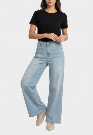 Mujer con camiseta negra de manga corta, jeans anchos de talle alto azul claro y zapatillas beige, de pie con las manos entrelazadas.