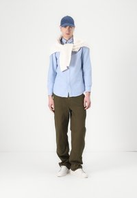 Lichtblauw overhemd, olijfgroene broek, crèmekleurige sweater over de schouders gedrapeerd, blauwe pet en witte sneakers; gladde texturen zichtbaar.