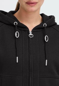 Crna zip-up hoodie s kapuljačom na vezanje. Ima sivi hardver, teksturiranu tkaninu i kengur džepove. Zaslon blizu područja vrata.