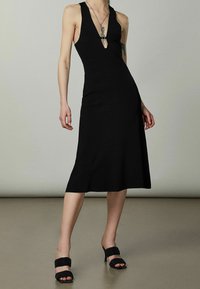 Abito midi nero aderente con scollatura a V profonda, texture a coste e orlo svasato, abbinato a sandali neri con tacco alto.