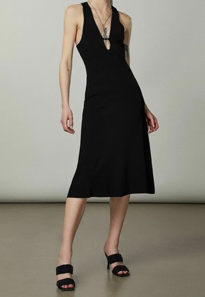 Robe midi noire ajustée avec un décolleté en V profond, texture côtelée et un ourlet évasé, associée à des sandales à talons hauts noires.