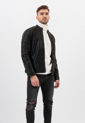 Black leather jacket with a stand collar, textured shoulder accents, worn over a white knitted turtleneck, paired with distressed black jeans.