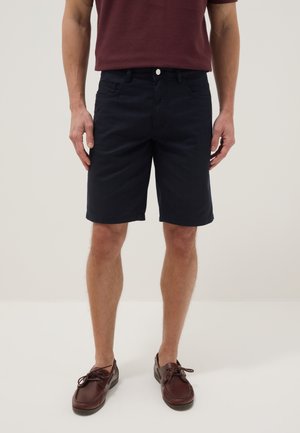 Mann trägt dunkelblaue Shorts, bordeauxfarbenes Hemd und braune Lederschnürschuhe, steht vor einem schlichten hellen Hintergrund.
