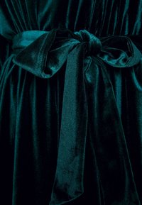 Teal sammetstyg med ett brett bälte knutet i en rosett. Texturen är mjuk och slät, med en lätt lyster och draperade veck.