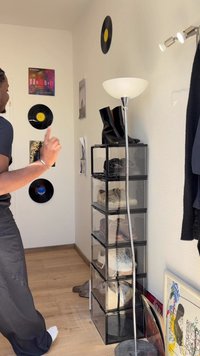 Unità di stoccaggio in acrilico trasparente che contiene vari scarpe, sormontata da stivali neri fino al ginocchio. Il muro espone dischi in vinile e manifesti. La lampada è in metallo.
