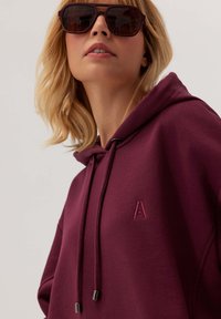 Bordowa bluza z kapturem, sznurkami i logo 'A'; wykonana z miękkiego materiału, w luźnym kroju. Model nosi ciemne okulary przeciwsłoneczne.
