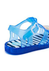 Sandalia de plástico transparente azul con hebilla, que presenta una plantilla a rayas. El diseño cuenta con recortes para una mayor transpirabilidad y una suela plana.