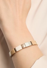 Pulsera de tono dorado con eslabones rectangulares entrelazados en colores beige, blanco y azul claro, que presenta un detalle redondo con el logo.