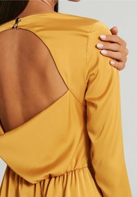 Gul satin klänning med figurnära midja, långa ärmar och en öppen ryggdesign. Har en liten knappdetalj vid halsringningen.