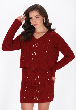 Kvinde med langt bølget hår iført en rød kabelstrikket sweater og nederdel med perleudsmykninger, poserer med den ene hånd nær hovedet.
